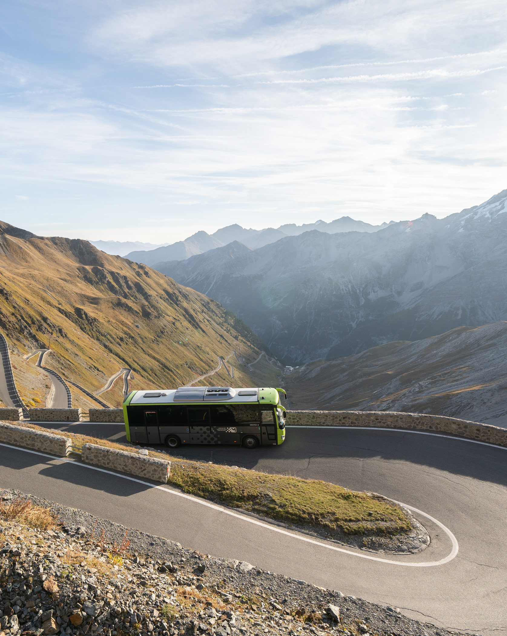 Ein grüner Bus fährt auf einer kurvenreichen Bergstraße mit malerischen Gipfeln im Hintergrund. - Demo-Seite